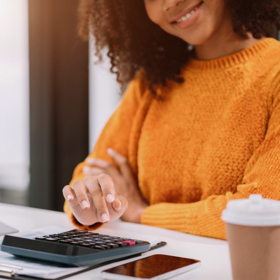 Smiling woman in an orange sweater using a calculator at a desk with a coffee cup.